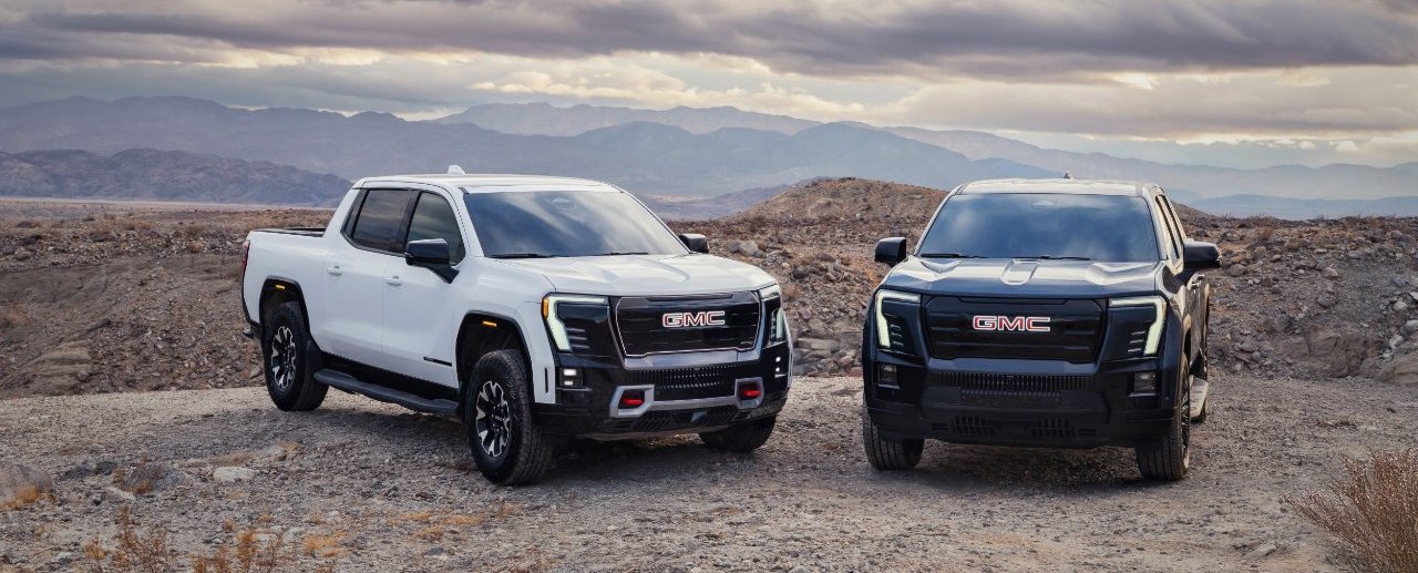 Pack view of the 2026 GMC Sierra EV including the AT4 (left) in Summit White, and Elevation (right) in Deep Ocean Metallic.